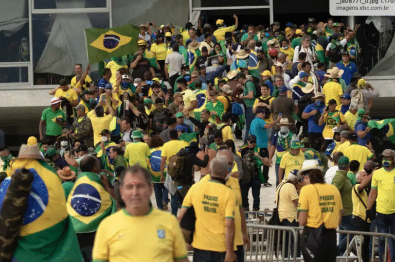 Uma grande multidão de manifestantes vestindo as cores verde e amarelo, as cores da bandeira brasileira, está concentrada em frente a um prédio público em Brasília. A imagem captura a densidade e o movimento do grupo.