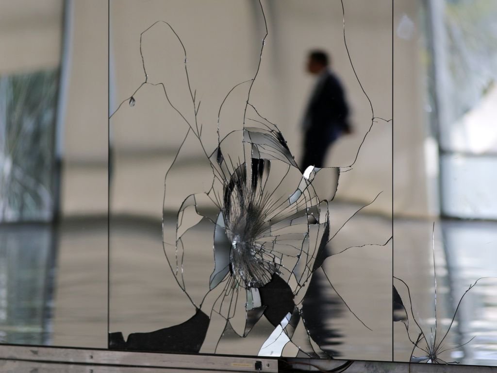 Fotografia em plano médio mostrando painéis de espelho quebrados em primeiro plano, com um grande ponto de impacto central e rachaduras radiais. No reflexo distorcido à direita, vê-se a silhueta desfocada de um homem de terno e gravata, afastando-se. O ambiente é um hall moderno e amplo com piso polido.